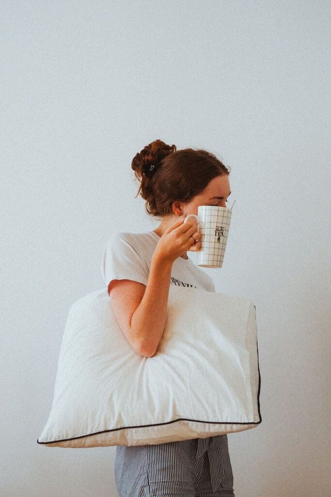 a woman holding a coffee mug that covers her face with a pillow under her arm