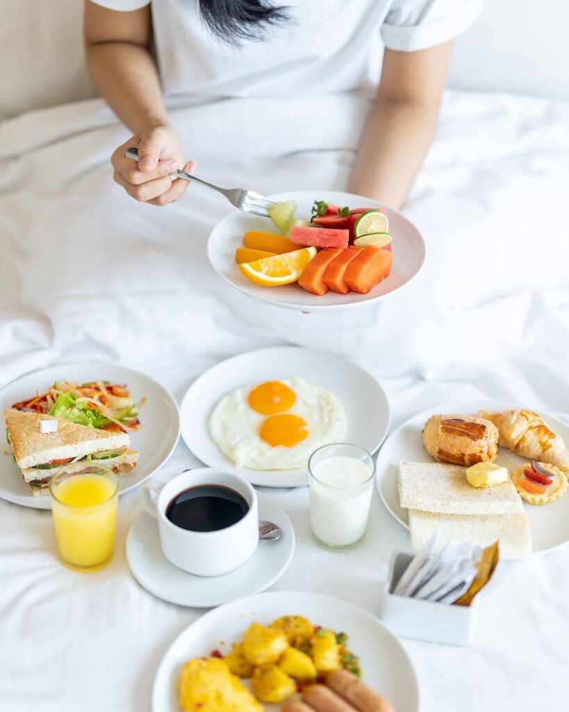 woman having a full breakfast at bed with fruit, bakery, coffee, milk, orange juice and sandwich