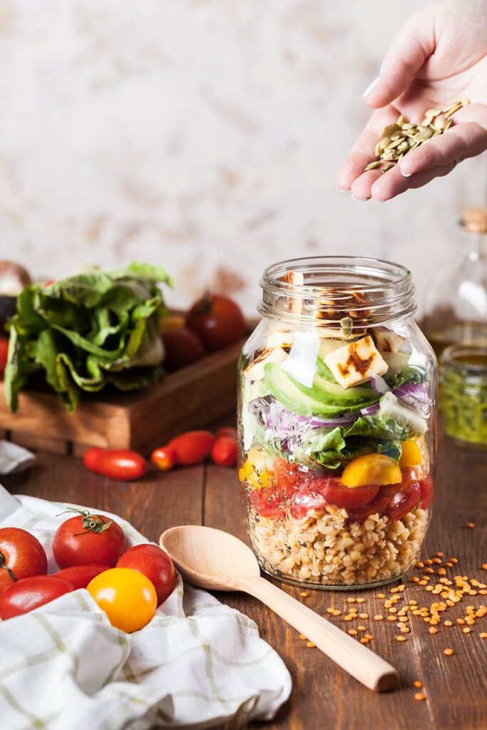 a jar containing a salad made of tofu, veggies, and lentils, veggies in the background and a wooden spoon lying on the table