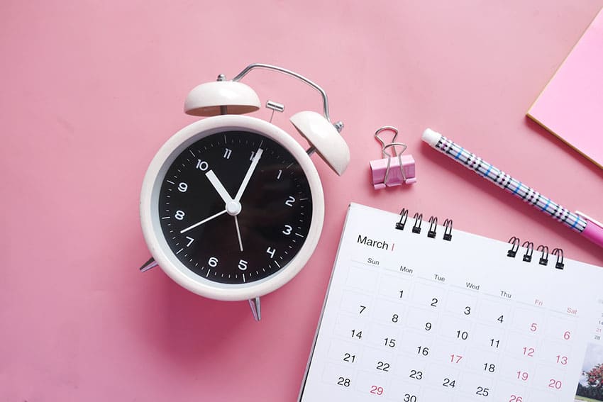 an alarm clock, a calendar and a pen over a pink surface