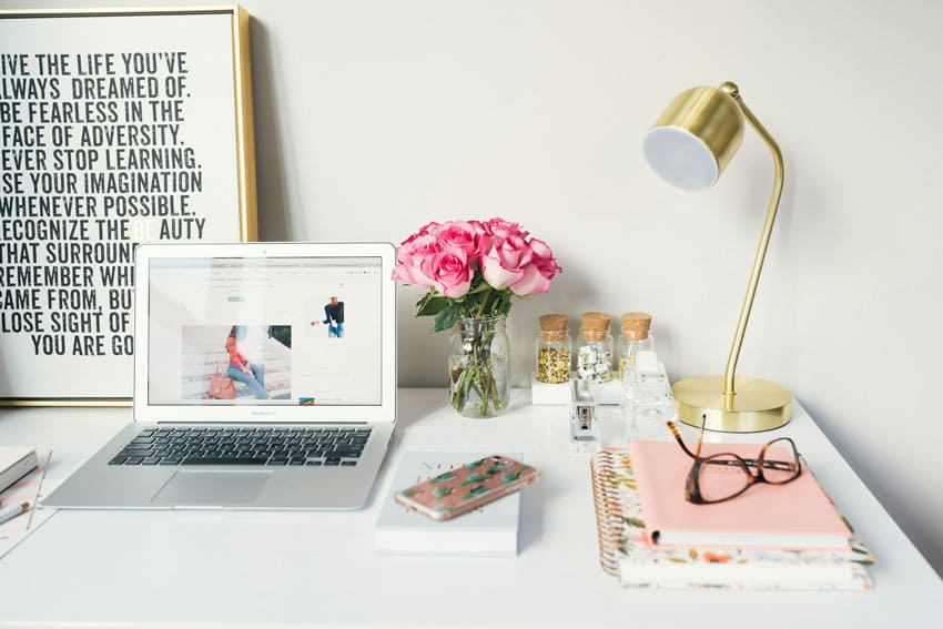 an office desk with lamp, books, a laptop, a print over the wall and a flower bouquet