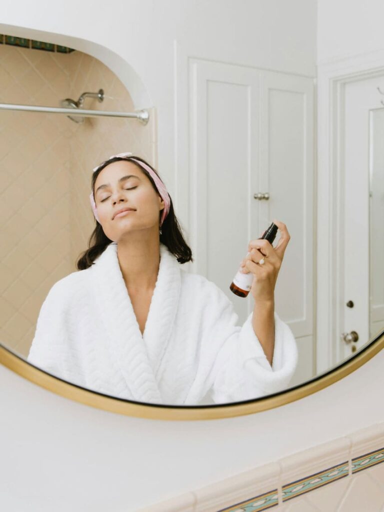 a brunette woman wearing a bathrobe in her bathroom and putting a soothing spray on her neck soothing spray seller as one of the best beauty side hustles