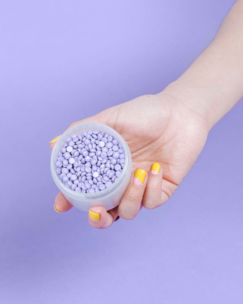 a woman's hand holding a container with purple wax beads over a purple background wax esthetician as one of the best beauty side hustles