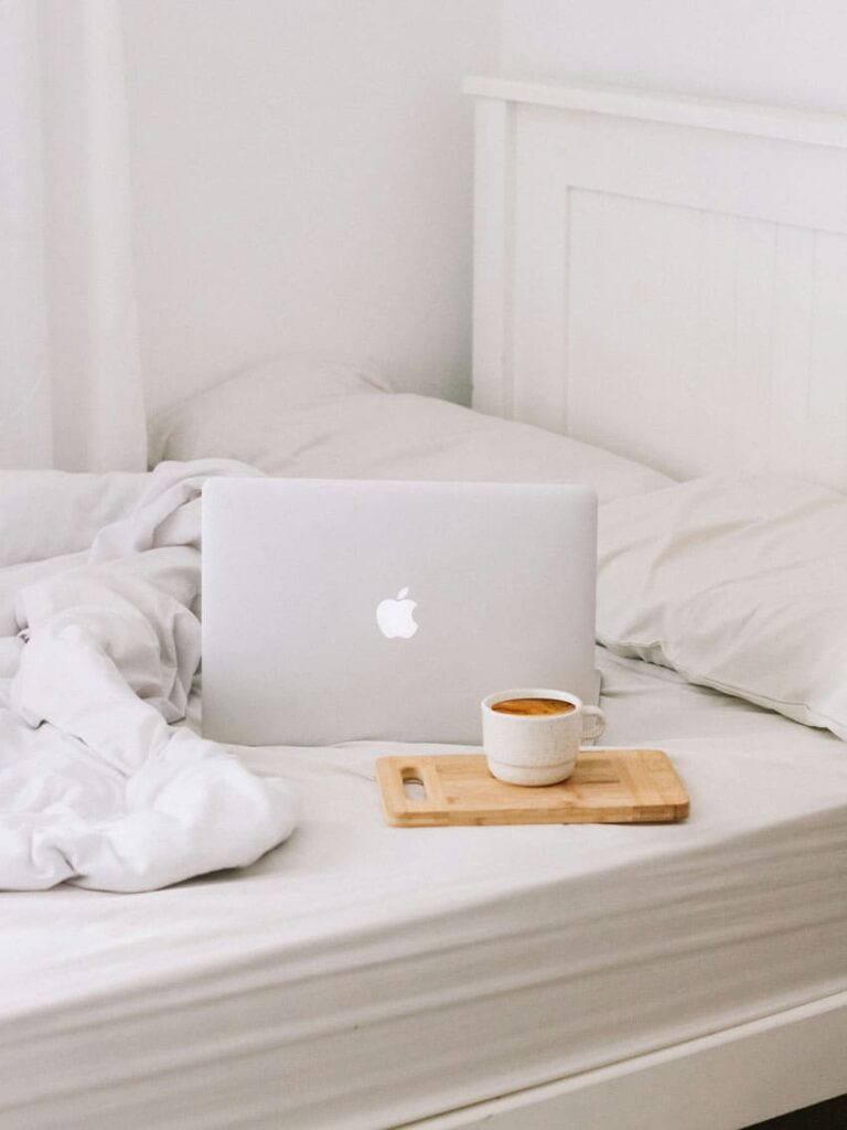 a white laptop over a bed, next to it there's a cup of coffee printable seller as one of the best beauty side hustles