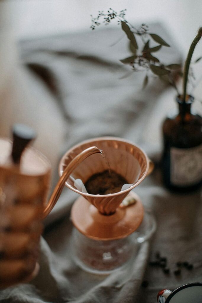 a woman pouring hot water from a copper kettle into a copper coffee maker